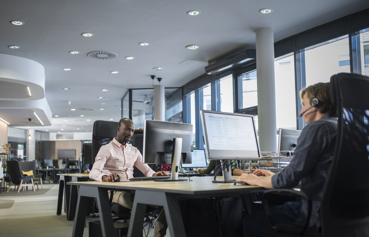 Two professionals working in a modern office environment, one wearing a headset and focused on a computer screen, the other engaged with a desktop monitor, illustrating business technology solutions and collaboration.